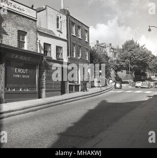1950s, historical picture looking up Heath Street, Hampstead with traditional shops of the day, such as E. W. Rout, family grocer. This picture was most likely taken on a s Sunday as the shop is closed and the road with litte traffic.  Hampstead is now promoted as the 'orginal urban village'. Stock Photo