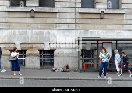 Homeless man sleeping on the ground next to a London bus shelter being ignored by waiting people outside Waterloo station London England UK Stock Photo