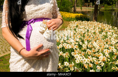 Pregnant woman is waiting her baby. This photograph is taking while she was holding shoes in her hand in front of flowers and a lake. Stock Photo
