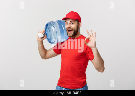 Portrait smiling bottled water delivery courier in red t-shirt and cap carrying tank of fresh drink and showing ok isolated over white background. Stock Photo