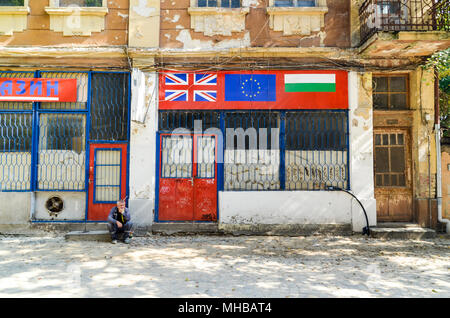 Old man sitting by a street, in front of a shop with the Bulgarian, UK and EU flags. Plovdiv, Bulgaria