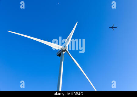 Plane flying behind a wind turbine on a blue sky background Stock Photo