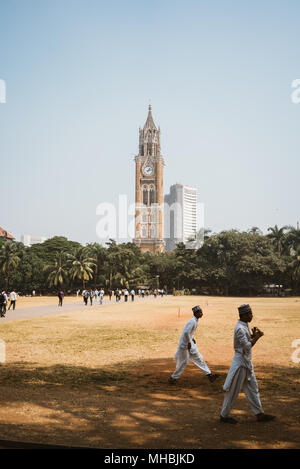 Indian boys playing cricket in a dry field with a gothic clock tower in the distance Mumbai India Stock Photo