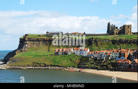 Whitby Abbey on a grassy headland as seen across the River Esk from Whitby town, with a row of red roofed houses and a sandy  beach in the foreground Stock Photo
