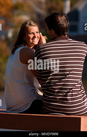 A young man gently caresses the smiling, sunlit face of his girlfriend while sitting on a bench. Stock Photo