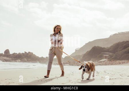 Full length shot of mature woman taking her dog on walk on the beach. Senior female on morning walk with her pet dog.