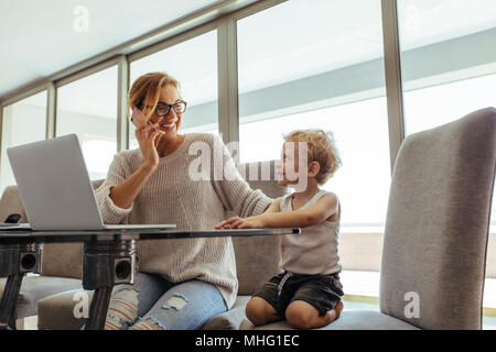 Young mother with son working from home, talking on the phone and smiling. Busy young woman with son working in home office. Stock Photo