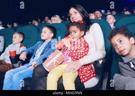 Beautiful woman and pretty little girl posing in movie theatre. Girl sipping fizzy drink and holding popcorn paper bag. Small spectators sitting in comfortable chairs, watching at screen and smiling. Stock Photo