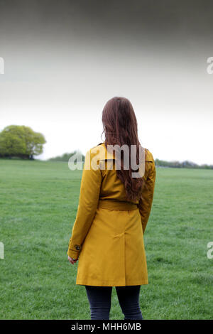 Young woman standing in a park with her back to camera looking at the horizon, she is wearing a ochre raincoat. Stock Photo