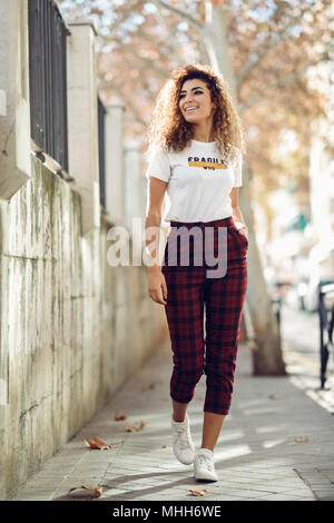 Beautiful young arabic woman with black curly hairstyle. Arab girl in casual clothes in the street. Happy female wearing white t-shirt and checked pan Stock Photo