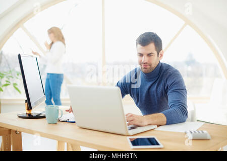 Portrait of handsome young businessman wearing casual clothes while sitting at laptop and typing. Uncrecognizable businesswoman standing at the backgr Stock Photo