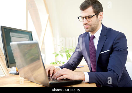 Portrait of young businessman wearing suit while sitting at desk in front of laptop and typing on keyboard. Stock Photo