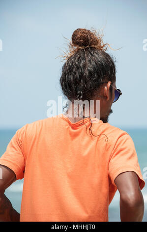 Flag Rock, Galle, Sri Lanka: Portrait (rear view) of a man with long curly hair, fashionably tied on the top of his head, standing looking out to sea Stock Photo