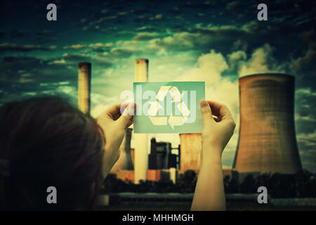 Woman holding a paper sheet with recycle symbol over factory pipe polluting air background, with smoke from chimneys going up in the sky. Environmenta Stock Photo