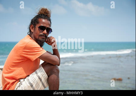 Flag Rock, Galle, Sri Lanka: Portrait of a man posing, head resting on hand, against a blue ocean background. Stock Photo