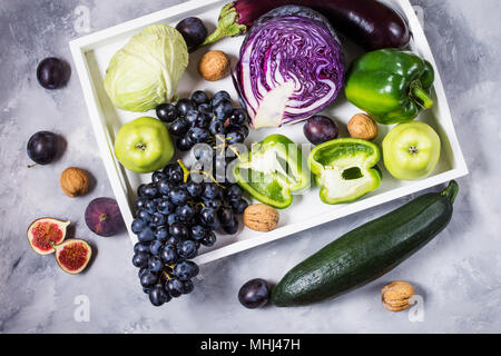 Fresh organic raw green and purple colored vegetables and fruits in white tray on dark stone background. Stock Photo