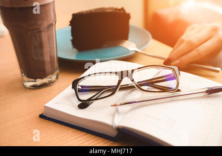 Still life: Eyeglasses and pen on open notebook.  Coffee cup and piece of cake. Fork in hand. Planning concept Stock Photo