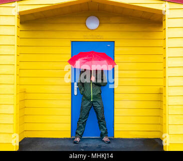Saltburn by the sea, North Yorkshire, England, UK. 2nd May, 2018. Weather:  A splash of colour on a wet and windy  Wednesday morning on the Yorkshire coast as a walker shelters under colourful beach huts overlooking Saltburn beach. A band of rain is forecast to sweep across many parts of the UK on Wednesday morning, clearing to the east. Dryer and warmer weather is forecast for the approaching Bank Holiday weekend. Credit: ALAN DAWSON/Alamy Live News Stock Photo