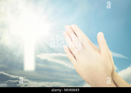 Concept of Christian prayer - folded hands for prayer against a blue sky and a shining cross sign (mixed) Stock Photo