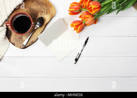 Overhead shot a bouquet of orange and yellow tulip flowers with black coffee, empty card with copy space, and a cozy knit throw blanket over white woo Stock Photo