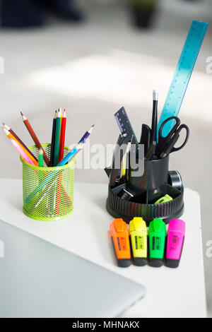 On the desk in the interior of the apartment are stationery items. Multicolored pencils, markers and globe. On the table is a closed laptop Stock Photo