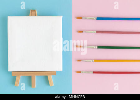 Flat lay of art canvas on easel and colorful paintbrushes on blue and pink background minimal creative concept Stock Photo