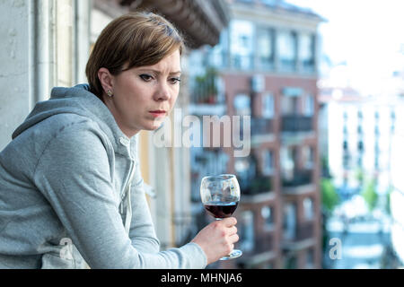 Beautiful red haired caucasian woman suffering from depression holding a glass of wine on a balcony at home. Staring out feeling sad, pain and grief.  Stock Photo