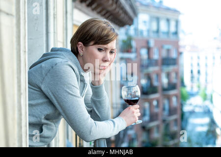 Beautiful red haired caucasian woman suffering from depression holding a glass of wine on a balcony at home. Staring out feeling sad, pain and grief.  Stock Photo