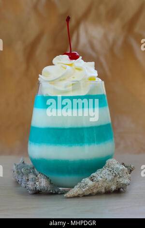 Blue striped jello dessert in glass with whipped cream and red candied cherry on top, seashells, sea vacation concept Stock Photo