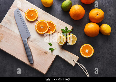Flat lay of fresh citrus fruits, half cut orange and lemons on cutting board with knife on black stone background. Horizontal top view. Stock Photo