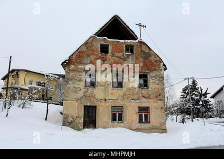 Abandoned old ruined house with broken windows, fallen facade and bricks from walls, covered in deep snow next to paved road and with modern suburban Stock Photo