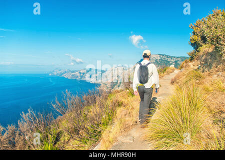 young caucasian man wearing white shirt, cap and backpack walking along Path of Gods trail on Amalfi coast on sunny summer day, Salerno, Campania, Ita Stock Photo