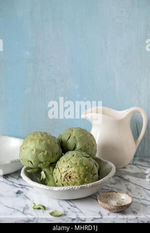 three artichokes ready in a white ceramic bowl with pitcher. Stock Photo