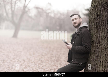 A smiling handsome young hipster man leaning against a tree and checking his smartphone in the park. Stock Photo