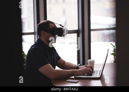 Male executive using virtual reality headset with laptop at desk Stock Photo