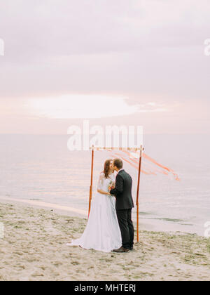 The kissing newlyweds during the wedding ceremony on the beach at the background of the sunset. Stock Photo
