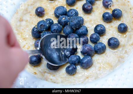 Person hand holding spoon scooping up breakfast porridge with blue berries and honey on top on a white plate Stock Photo