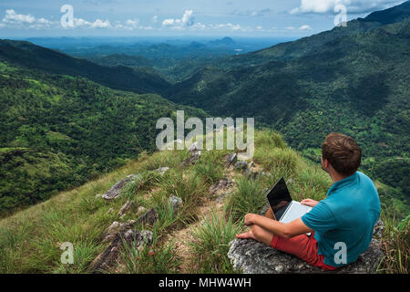 caucasian man with laptop sitting on the edge of ella mountain with stunning views of the valley in Sri Lanka. Stock Photo