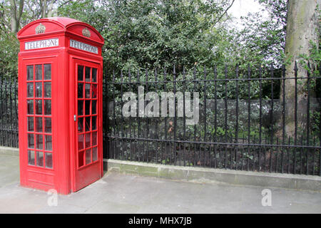 Red London Telephone Box, on pavement by fence outside a park Stock Photo
