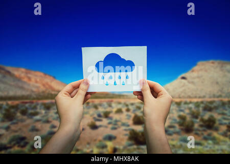 Close up of woman hands holding a white paper sheet with a rain cloud icon over a hot desert background. Dreaming of a rainy day in a sunny season. Stock Photo