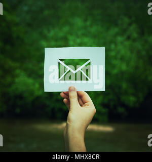 Woman hand holding a paper sheet with envelope message icon over green forest nature background. Ecology and greening concept, global environmental is Stock Photo