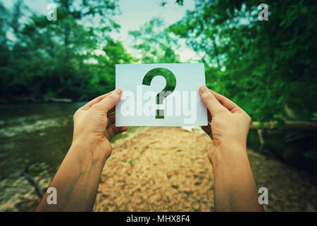 Close up of woman hands holding a paper sheet with the question mark inside, over green forest nature background. Lost people concept, global environm Stock Photo