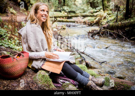 Young woman relaxing on forest riverbank with notebook Stock Photo