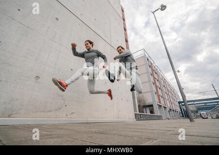 Young adult male twin runners, running and jumping mid air on city sidewalk Stock Photo