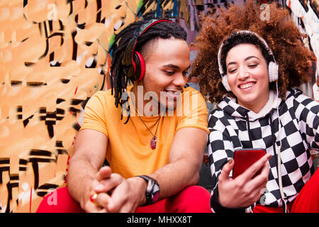 Two friends wearing headphones, listening to music, holding smartphone Stock Photo