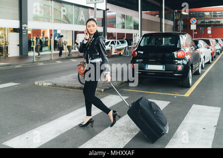 Woman walking outdoors, pulling wheeled suitcase, talking on smartphone Stock Photo