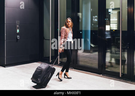 Woman walking towards hotel, pulling wheeled suitcase, looking over shoulder Stock Photo