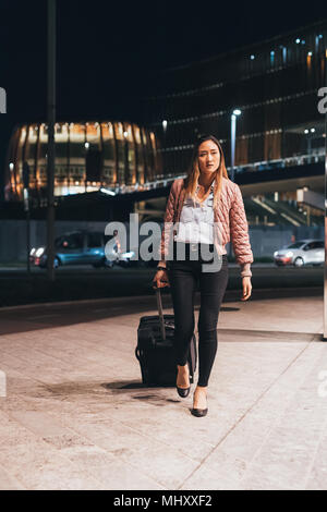 Woman walking outdoors at night, pulling wheeled suitcase Stock Photo