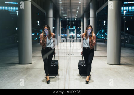 Woman walking beside modern building at night, pulling wheeled suitcase, reflection in window Stock Photo