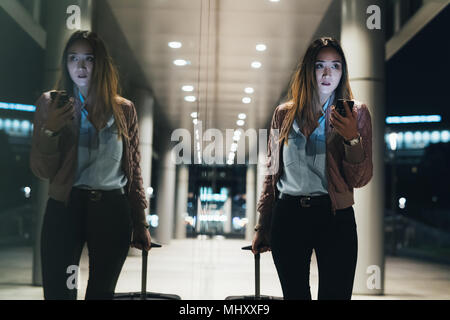 Woman walking beside modern building at night, pulling wheeled suitcase, reflection in window Stock Photo
